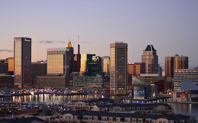 The sun sets over the Baltimore skyline. Tall building dominate the background with low rowhouses lining the foreground.