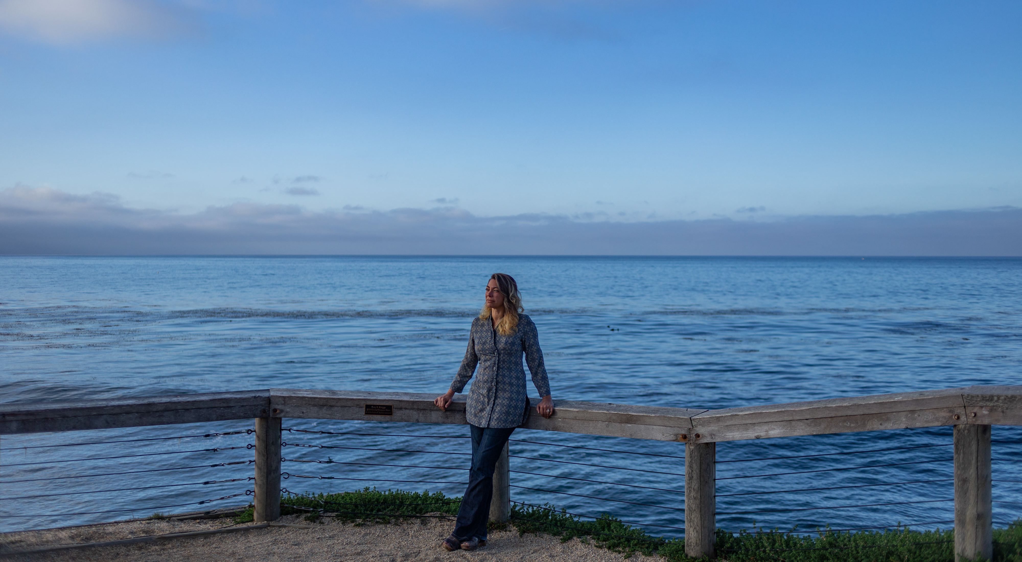 Former TNC lead scientist Heather Tallis leans against a railing facing the camera, with a vast blue Pacific Ocean horizon behind her.