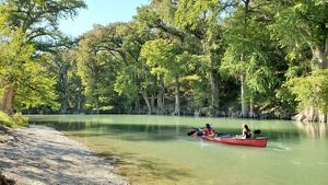 Two kids paddle a kayak on a blue river lined with dense trees.