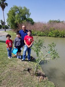 Three young boys stand with their father on the edge of a river beside a young Montezuma cypress tree that has just been planted.