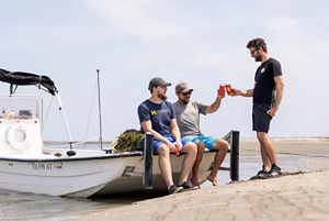 Two men sit on the stern of a small Carolina skiff pulled up on a beach. One seated man clinks cans in a cheers with a man standing on the beach.