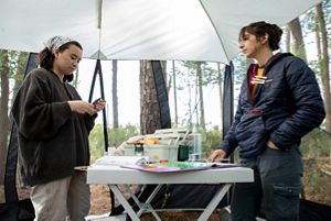 Two women stand across from each other with a long folding table between them. The table holds a number of materials and supplies needed for a bird banding project.