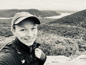 A person take a selfie in front of a vista view of a winding river curving around the base of a small mountain.