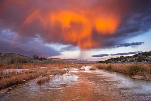 Virga falls from orange and purple clouds over a rocky creek.
