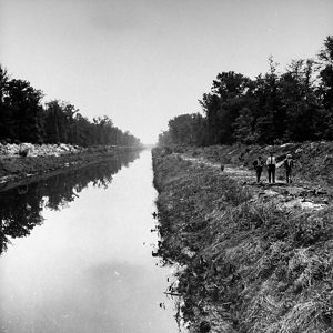 Black and white photo taken in 1943. Three men walk along a narrow river channel. The banks on either side are piled with soil forming high berms.