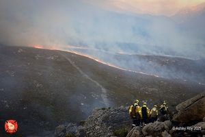 Four people wearing fire gear stand on a rocky ledge looking out toward lines of fire burning on hillsides in the distance.