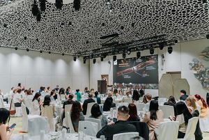 People sitting at round tables facing a speaker on a stage.