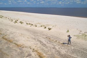 A man stands alone on a wide stretch of beach, using a long-handled tool to create holes to plant sea oats. The blue waters of the Gulf stretch behind him to the horizon.