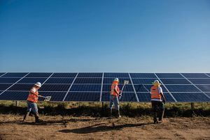 Three men in hard hats and orange utility vests clean large solar panels on a sunny day.