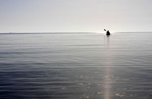 A person floats in a kayak in a flat, wide body of water. They are silhouetted against the sky and seem to hover in the distance at the horizon.