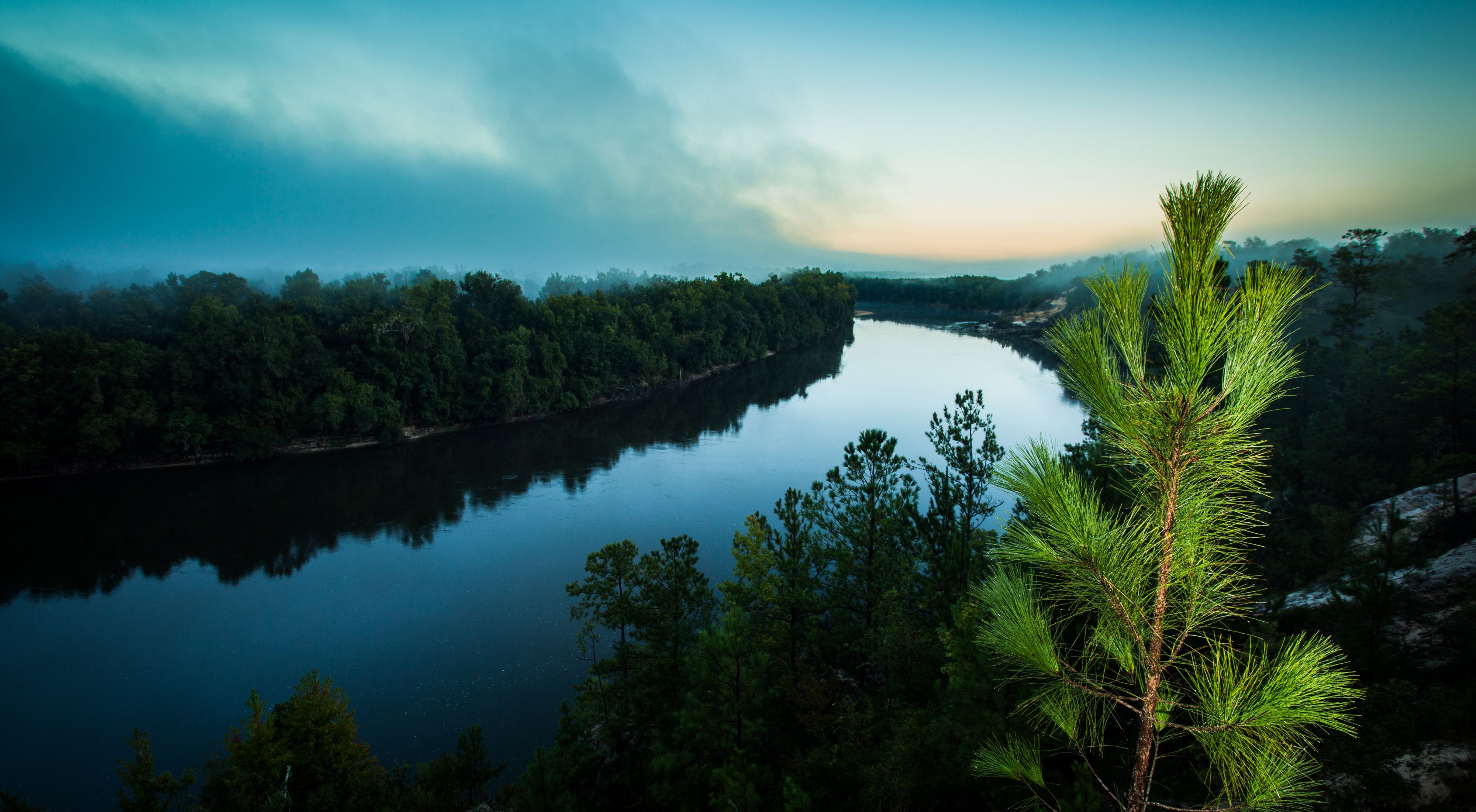 A wide river bends through a forested landscape with trees along the banks and a misty blue sunset.
