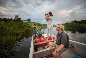 Three people on a boat on a body of water, one stands with binoculars looking out into a lush vegetated area.