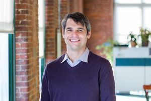 Portrait of Alex Mas as he smiles toward the camera with a blurred office space behind him.
