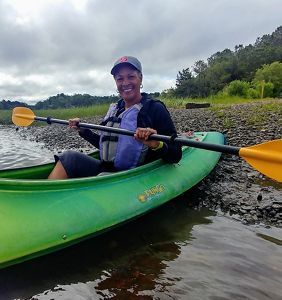 Dale Threatt-Taylor sostiene un remo dentro de un kayak, en una orilla frente a un paisaje boscoso.