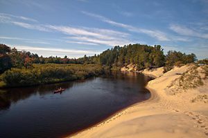 People kayak down a bend of the Two-Hearted River in Michigan. On the left of the river is autumn foliage and on the right is a sandy shore. 