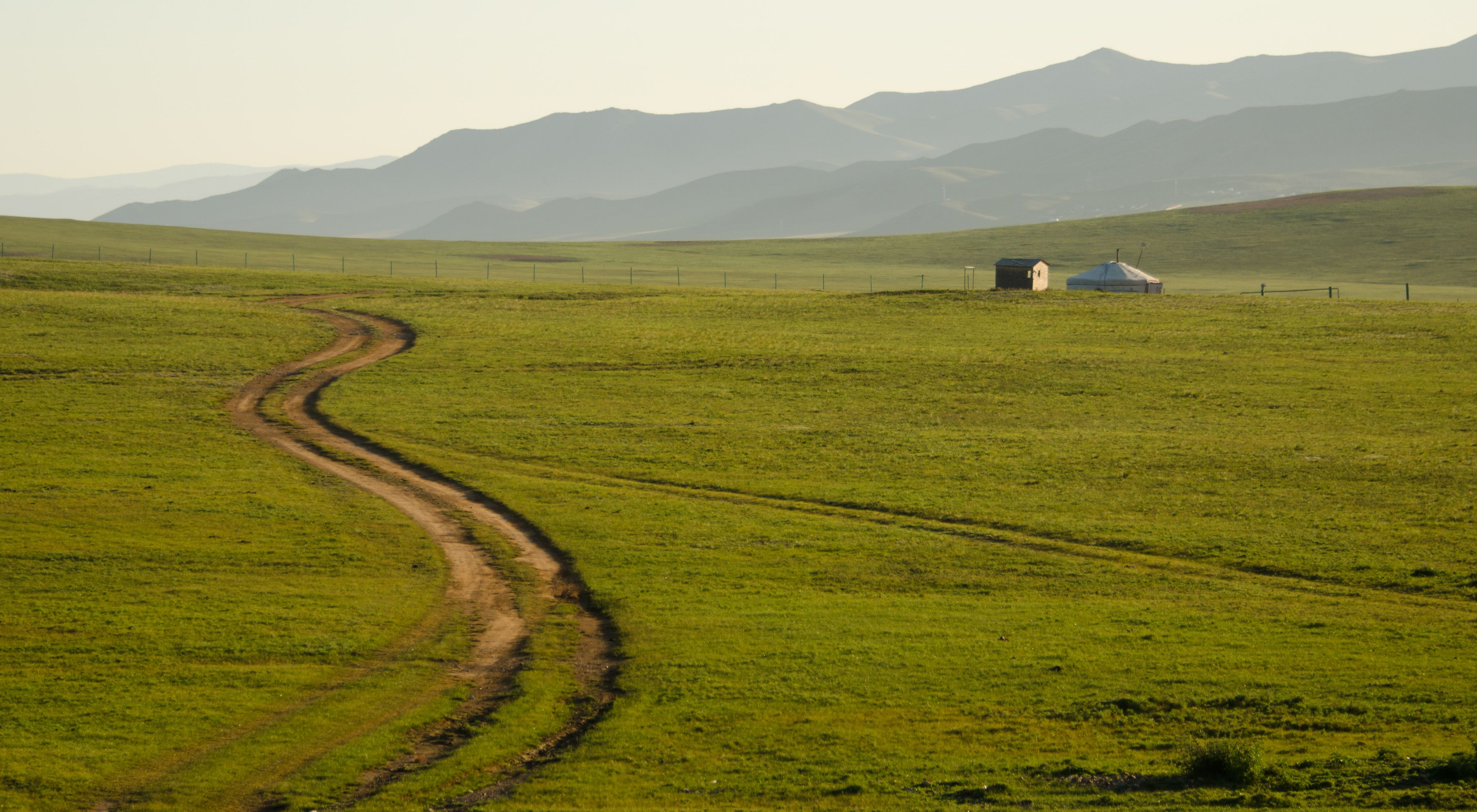 A dirt road winds through open green grassland with a yurt and an outbuilding in the middle ground and mountains in the far distance.