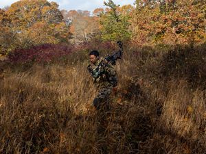 man in camo walks through grassy field holding compound bow.