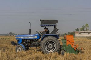 Amandeep drives through a dry rice field with a blue tractor with an orange attachment in back known as the super seeder. 