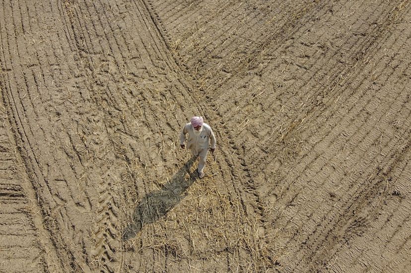 Aerial of Amar Singh walking over a dry bare farm field with tractor tracks that has just been seeded.
