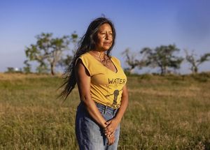 woman stands in yellow shirt, long hair, looking into the distance, brown and green grass and small trees behind her