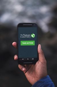 A hand holds a smartphone that has the TNC logo and the words 'Take Action' on the screen.