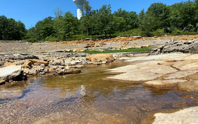 A stream runs through a rocky channel, with trees and a water tower in the background.