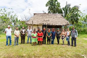 A group of people stand in a line in front of a small, grass-roofed building and trees.