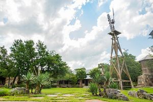 A windmill looms in front of a house surrounded by green grass and trees.