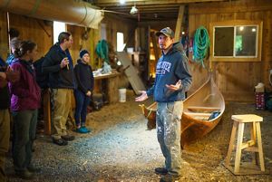 Michael Chilton, a Kasaan wood carver, stands in a wooden building with a dirt floor and two wooden canoes.