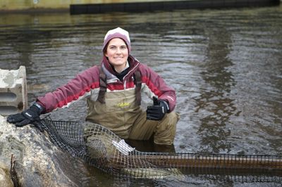 Woman in pink and red coat with gloves and a cap with chest waders sits in the water at the shorelines with restoration equipment in the foreground.