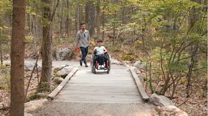 A boy using a wheelchair makes his way down a boardwalk together with his father.
