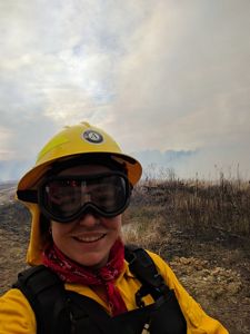 A woman wearing yellow fire retardant gear and thick goggles takes a selfie during a controlled burn. She is standing in front of an open scrubby piece of land that has not yet been ignited.
