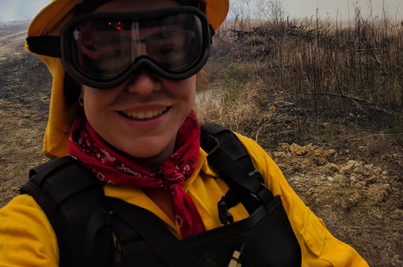 A woman wearing yellow fire retardant gear and thick goggles takes a selfie during a controlled burn. She is standing in front of an open scrubby piece of land that has not yet been ignited.