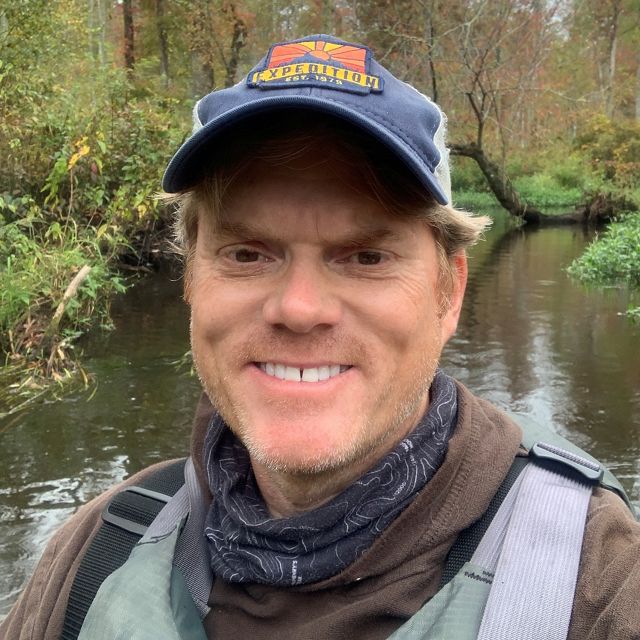 Headshot of a smiling man standing in a forested wetland.