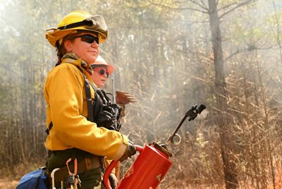 A woman wearing protective fire gear holds a large red drip torch canister during a controlled burn. White smoke rises in the air behind her.
