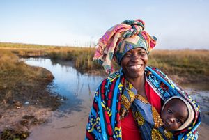 A smiling woman carries a child in a colorful fabric sling. She stands at the edge of a narrow creek.