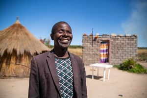 Candid portrait of village chief Fernando Kufuna. A smiling man stands outside with traditional village buildings in the background.