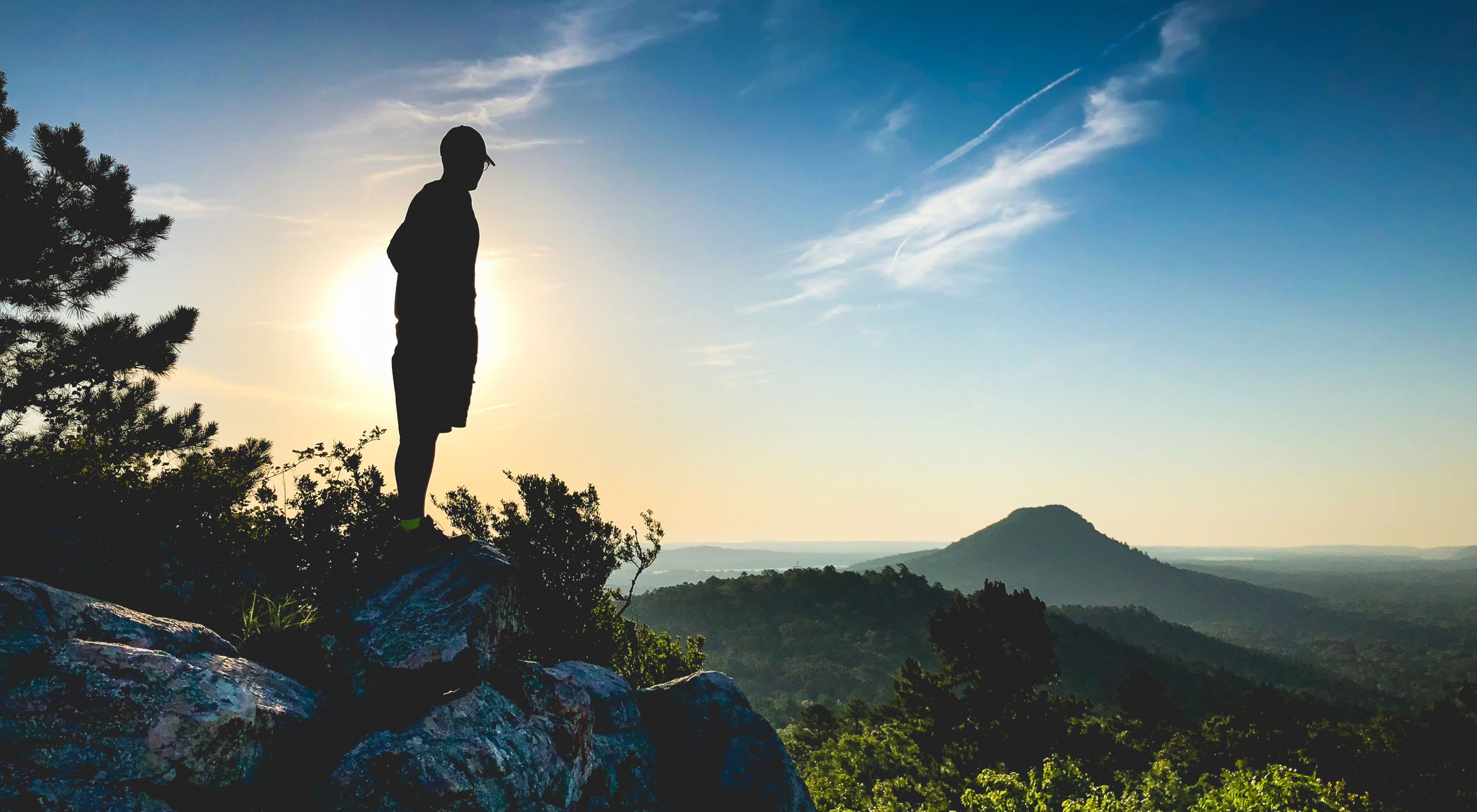 A hiker silhouetted against the sunset as he looks out over hills and valleys.