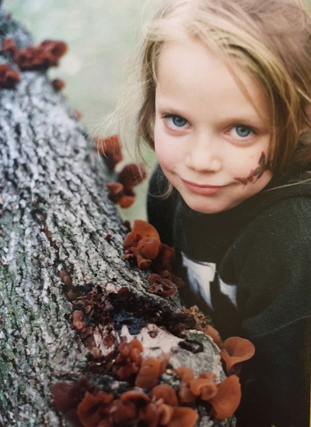 A little girl next to a tree trunk with reddish brown mushrooms growing on it.