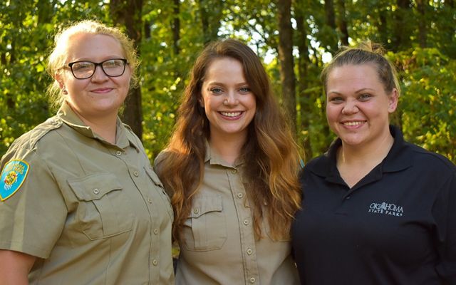 Three smiling women, two in khaki uniforms, with trees in the background.
