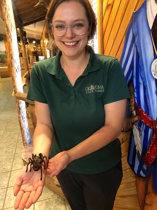 A young woman in a green, collared shirt, holding out a large spider on her wrist.