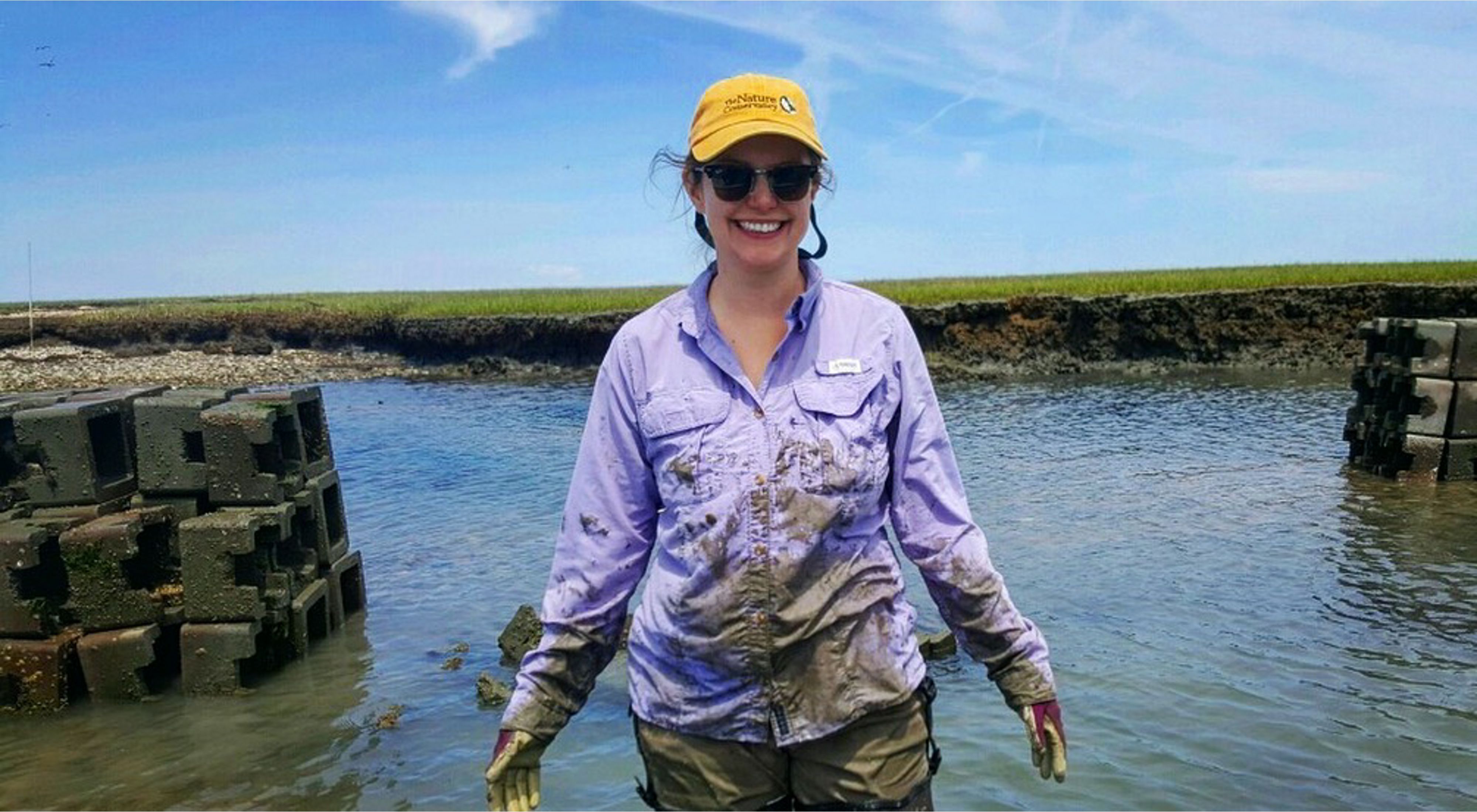 Angelina Stancampiano stands smiling in thigh-deep water with mud on her shirt and hands and oyster castles in the background.