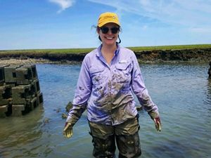 Angelina Stancampiano stands smiling in thigh-deep water with mud on her shirt and hands and oyster castles in the background.