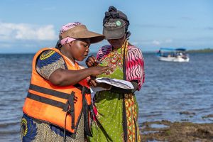 A woman, right, wearing a TNC baseball hat, stands on a beach and holds a notebook. Another woman, left, is pointing at something in the notebook.