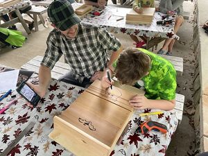 A man holds a picture of an owl up for a boy sketching the animal on a wooden box.