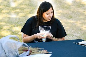 A woman sits at a table weaving with a bag filled with pine needles next to her.