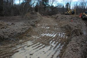 Standing water pools in the many depressions of a wide muddy channel created by a backhoe. 