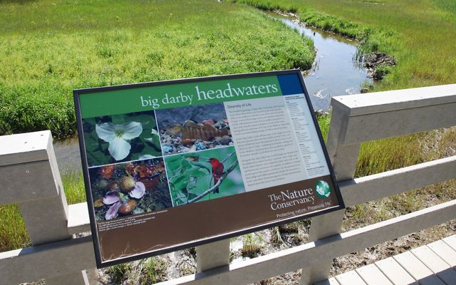 An interpretive sign attached to a wooden structure overlooks a meandering stream.