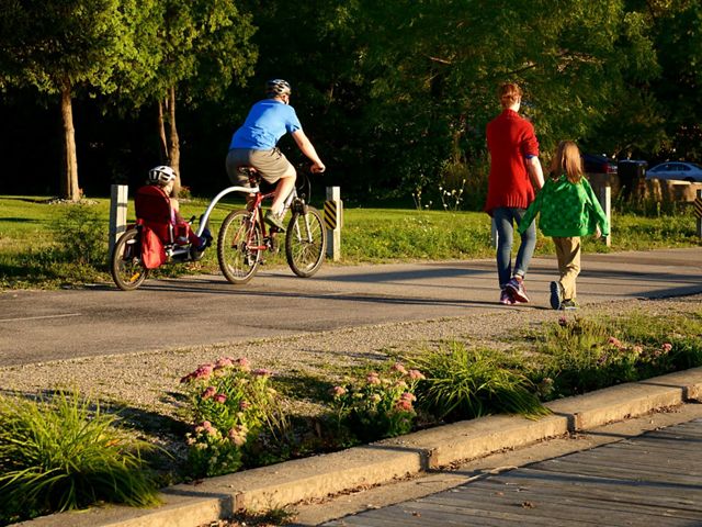 Man on a tandem bike with child in the back and woman with a second child walk along a trail bordered by planted pink flower