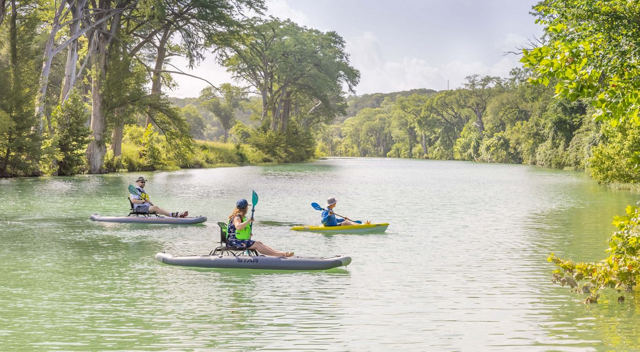 Three kayakers paddle on a blue river lined with trees.